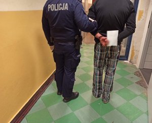 Na zdjęciu widoczny umundurowany policjant, który prowadzi zatrzymanego mężczyznę przez korytarz. Mężczyzna ma założone kajdanki na ręce trzymane z tyłu.