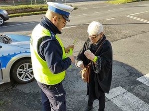 Policjant zakłada starszej kobiecie element odblaskowy na rękę przy przejściu dla pieszych.